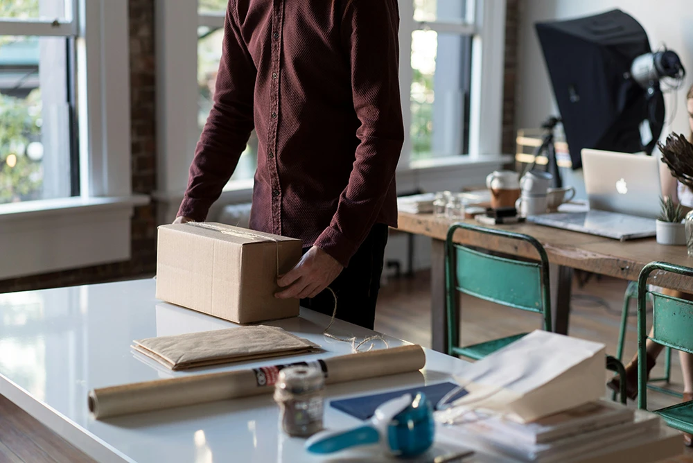 Eine Person verpackt ein Paket auf einem Schreibtisch in einem hellen Büro mit Laptop und Arbeitsmaterialien im Hintergrund.