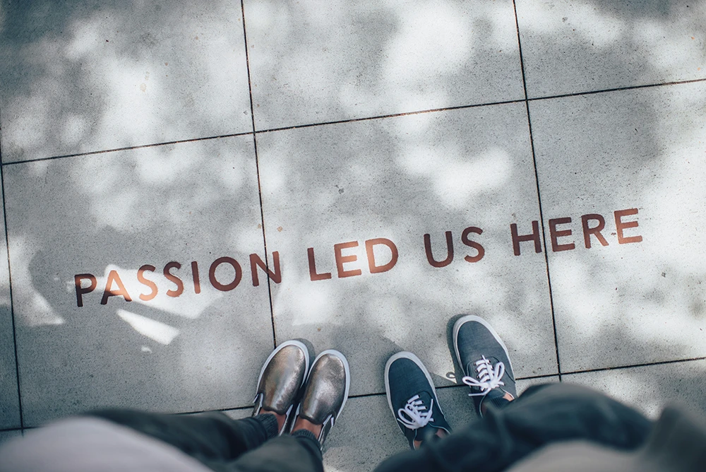 Zwei Personen stehen auf einem gefliesten Boden mit dem Schriftzug 'Passion Led Us Here' in Rot, umgeben von Baumschatten.