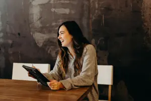 Lächelnde Frau mit Tablet im Gespräch an einem Holztisch vor dunkler Wand.