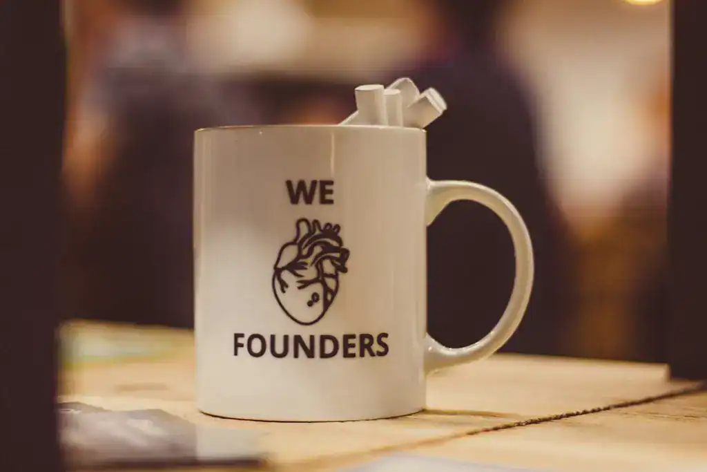 Weiße Kaffeetasse mit der Aufschrift We Founders und Herzgrafik auf Holztisch.