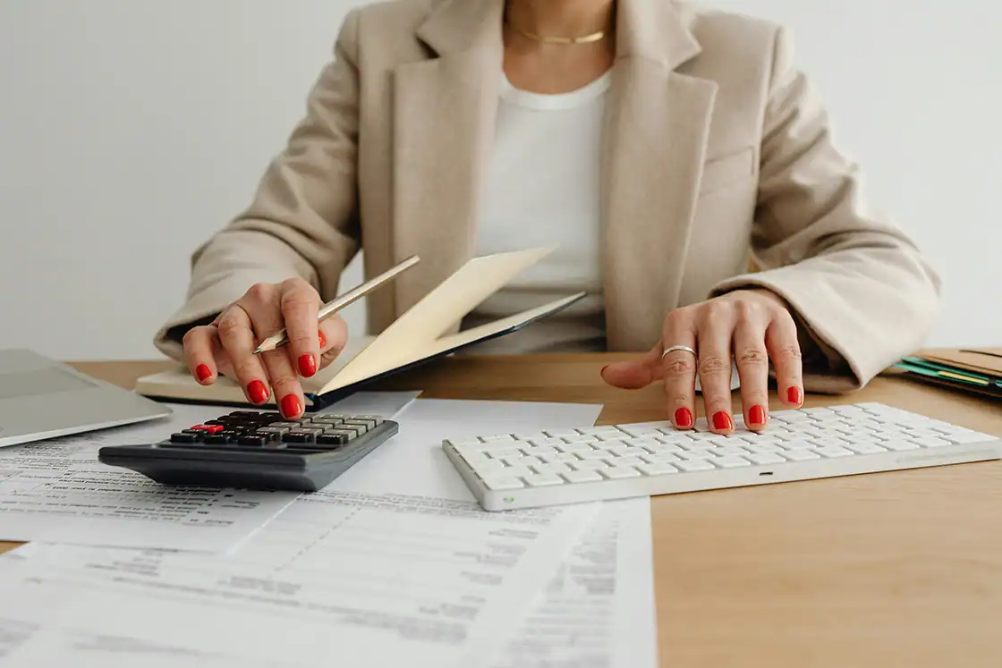 Frau arbeitet am Schreibtisch mit Taschenrechner, Tastatur und Unterlagen.