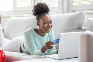 Frau liegt auf dem Sofa und bezahlt Online-Einkauf mit Kreditkarte am Laptop.
