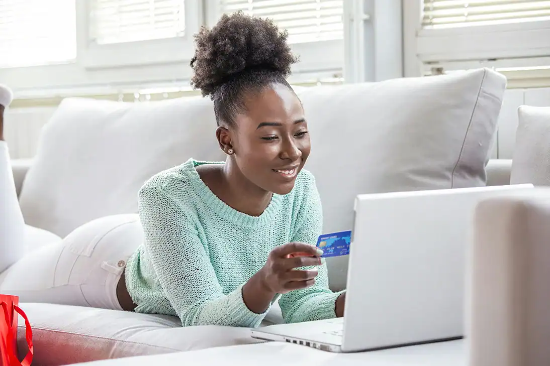 Frau liegt auf dem Sofa und bezahlt Online-Einkauf mit Kreditkarte am Laptop.