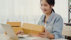 Frau sitzt am Laptop und hält ein Versandpaket in der Hand.