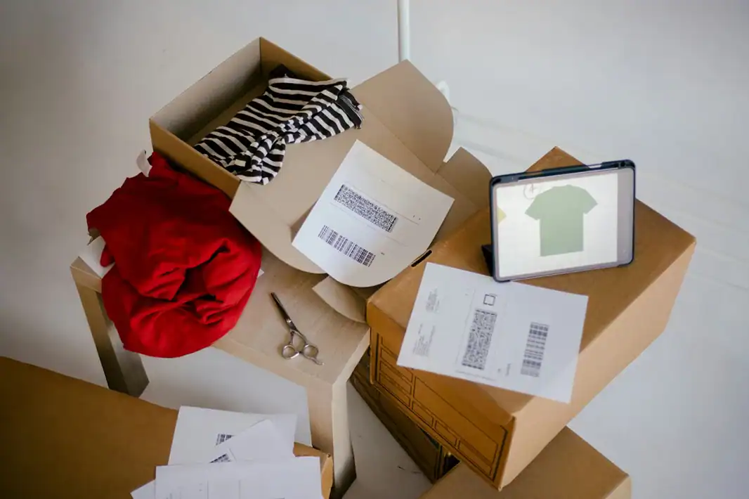 Versandkartons mit Kleidung, Etiketten und Tablet auf Holztisch